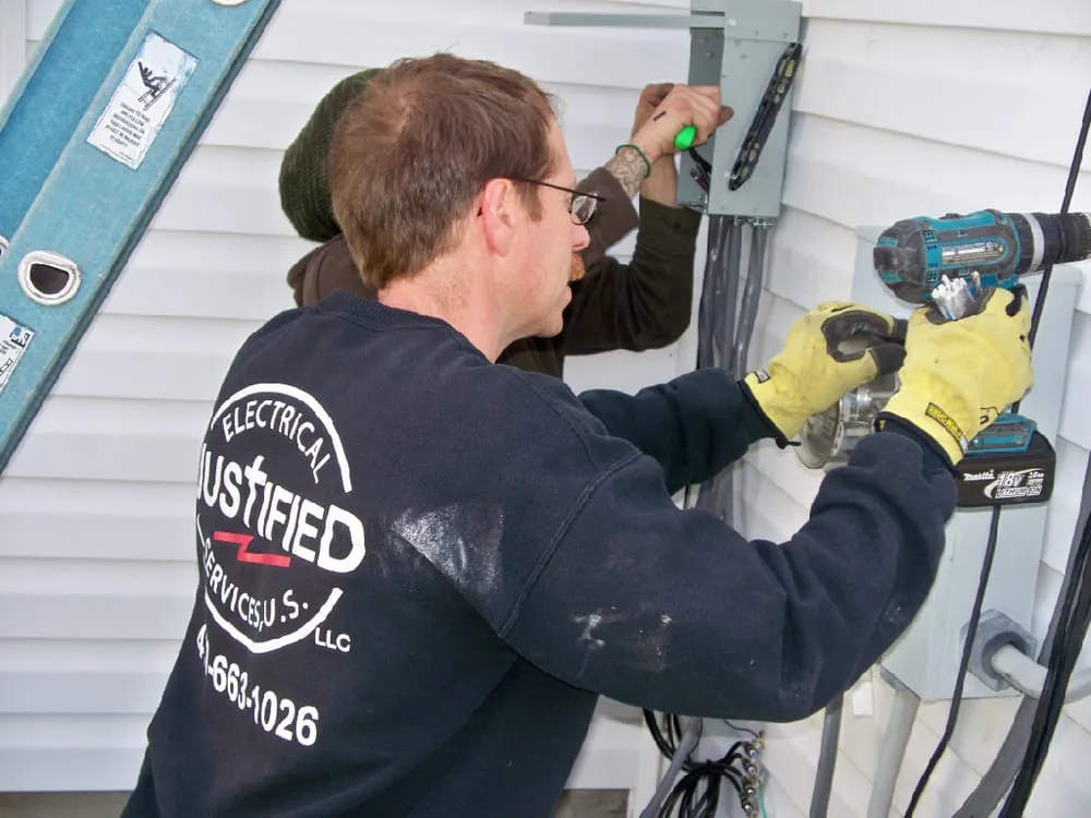 A Justified Electrical technician in a company sweatshirt drilling at the meter and service equipment on the side of a house