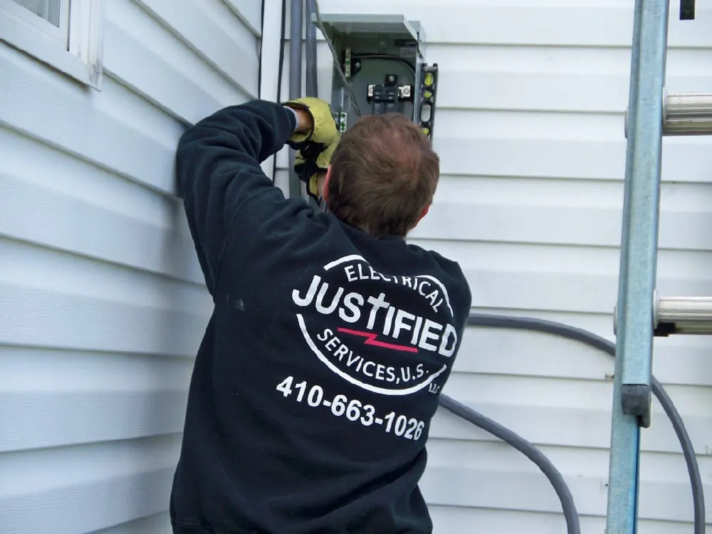 A Justified Electrical technician, back to the camera, working at an exterior panel beside a ladder